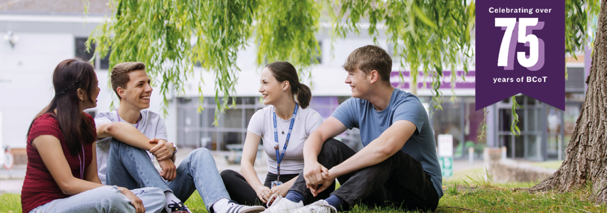 Students sitting outside the college on grass, under a tree
